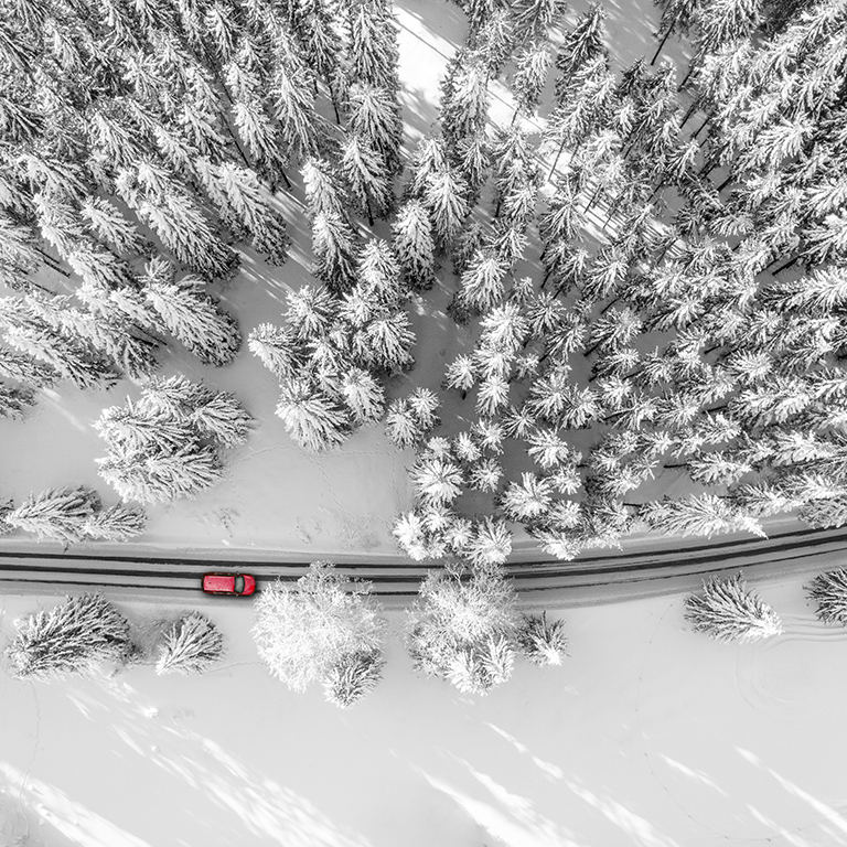 Road to nowhere. A drone view down to red car driving through a road between pine trees covered with snow.