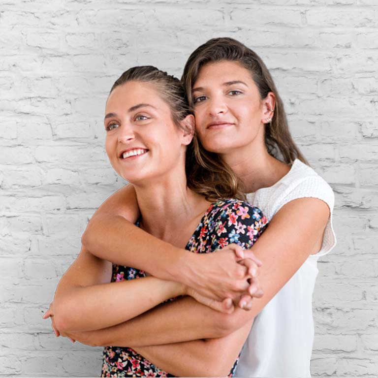 Portrait of smiling mid adult woman embracing young girlfriend. Happy lesbian couple is standing while holding hands. They are against white background.