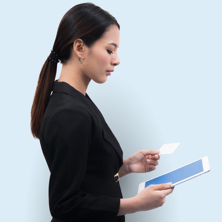 Young white woman dressed in black with a ponytail looks down to her tablet while she holds a business card. Her facial expresion is serious.