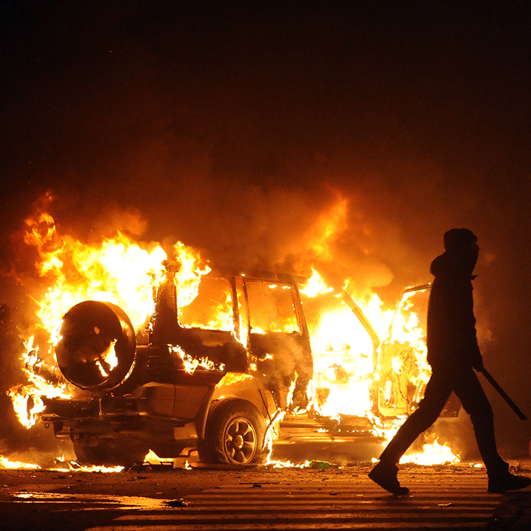 A dark figure walking away from a burning car at an unrest protest demonstration.