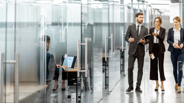 A team of three professionals is walking through a modern office space while discussing ideas. One member uses a laptop, showcasing a collaborative work environment.