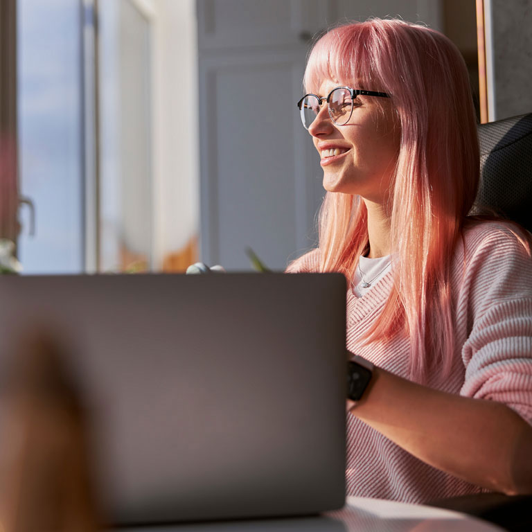 Gen Z pink haired woman with elegant glasses works on contemporary laptop near large window at home on sunny day