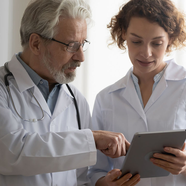 Medical team of two professional doctors talking and using digital tablet. Senior male chief physician helping young female nurse holding medical tech device discussing patient diagnosis in hospital.