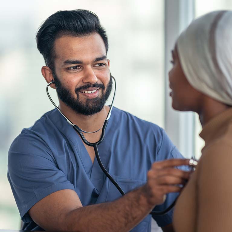 A mixed-race male doctor of Asian and Indian descent is performing a medical exam on a patient. The patient is a black woman with cancer. The woman is wearing a scarf on her head to hide her hair loss. The two individuals are seated next to each other. The medical professional is using a stethoscope to check his patient's heart and lungs.