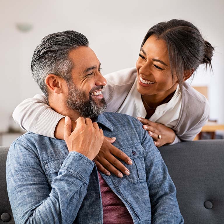Smiling ethnic woman hugging her husband on the couch from behind in the living room. Middle eastern man having fun with his beautiful young wife on the couch. MId adult indian man with latin woman laughing and looking at each other at home: complicity and love concept.