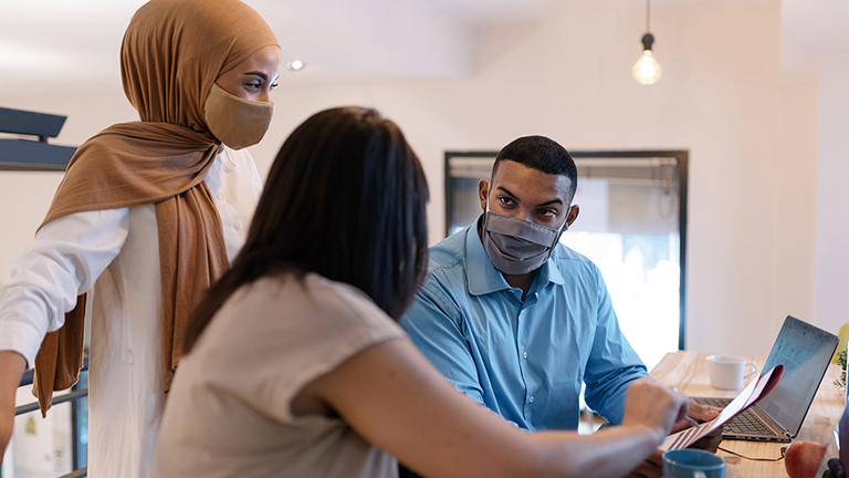 Stock photo of office workers wearing protective face masks due to covid-19 while working in the office. They are sharing ideas and using a laptop.