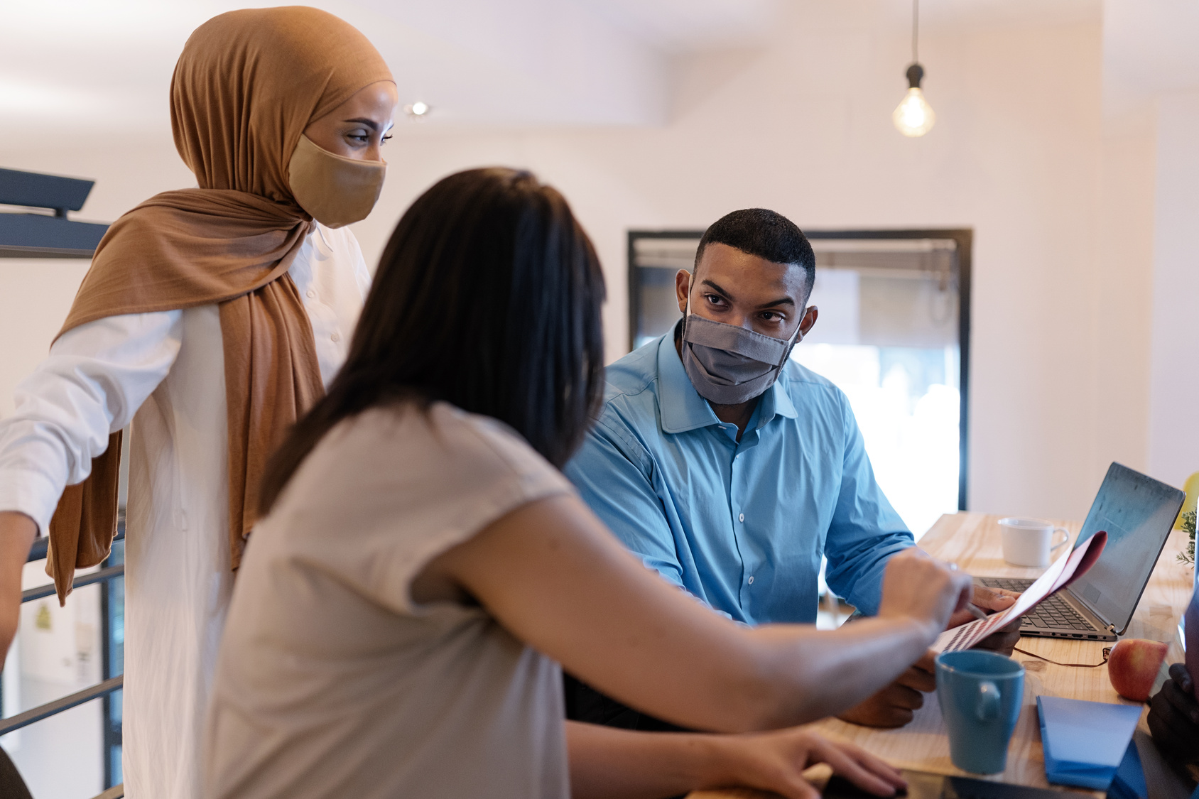 Stock photo of office workers wearing protective face masks due to covid-19 while working in the office. They are sharing ideas and using a laptop.