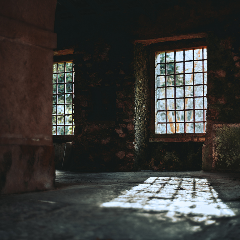 A dark atmospheric interior of an abandoned antique space with empty window holes with metal bars in them, a strong light spot from the window with a grid shadow on a stone ground, mossy grungy walls