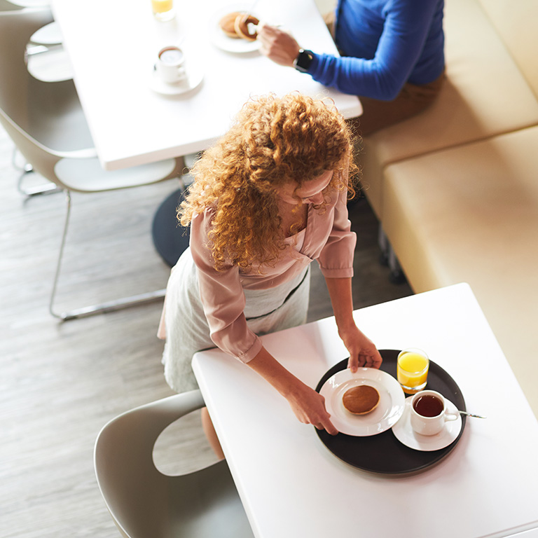 Aerial view of curly-haired young waitress standing at table and putting plate with pancake on tray