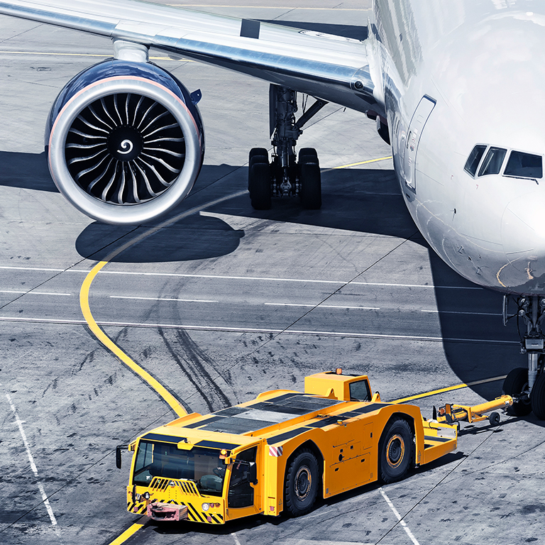 airplane on airport runway with pushback tractor attached to plane nose gear aerial top front view passenger jet engine aircraft towing by ground vehicle to terminal gate black and white wide banner