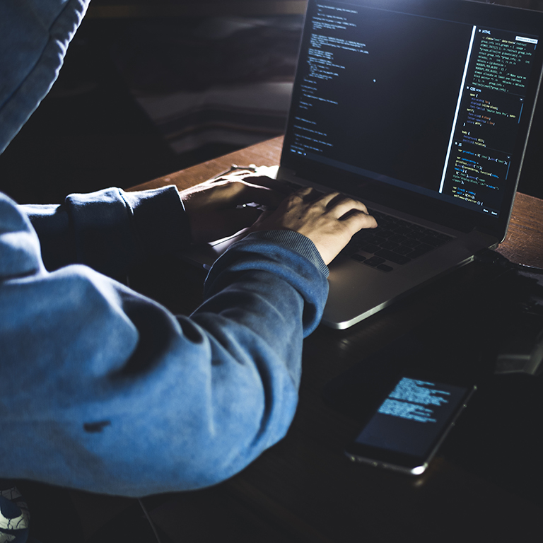 anonymous person in the hood sitting in front of computer working with laptop and mobile phone 