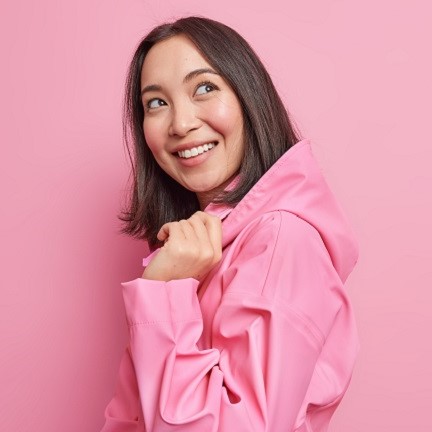 Pretty glad brunette Asian woman stands half turned against pink studio background has good mood wears stylish jacket with hood thinks about something pleasant poses happy indoor. Emotions concept