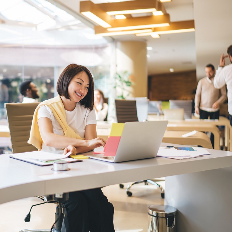 Ethnic young businesswoman in white tee shirt typing on laptop while sitting at office table in coworking space and smiling