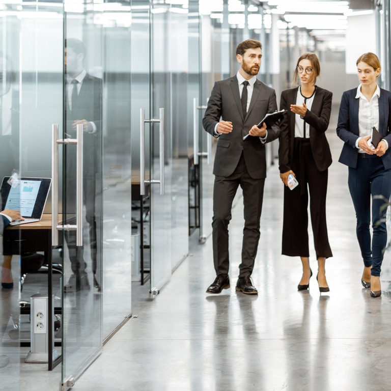 A team of three professionals is walking through a modern office space while discussing ideas. One member uses a laptop, showcasing a collaborative work environment.