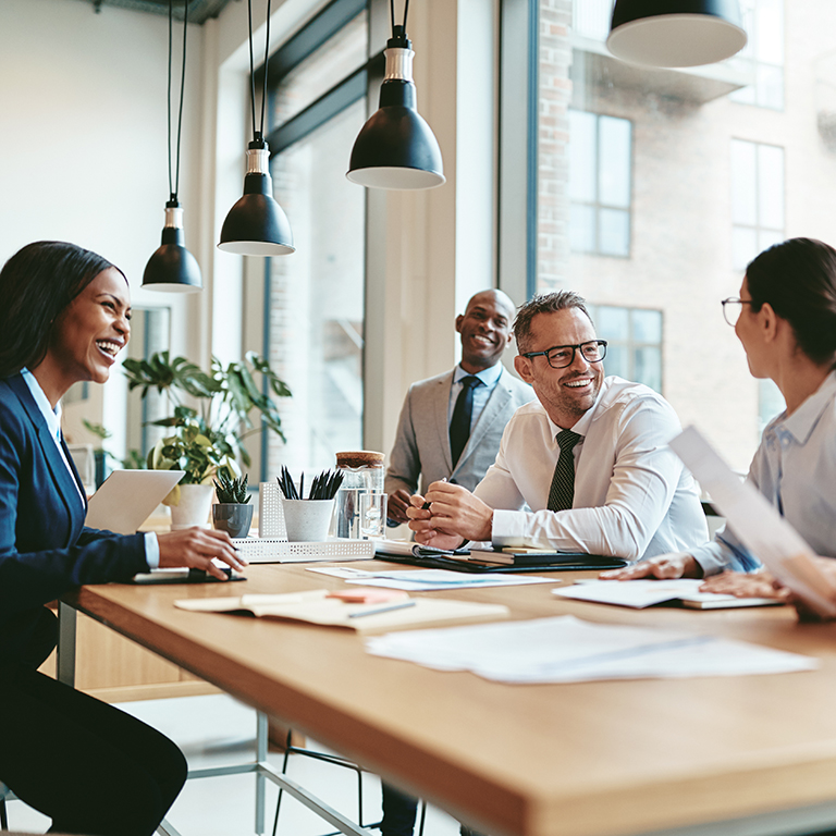 Diverse group of businesspeople laughing while discussing paperwork together during a meeting around a table in a modern office