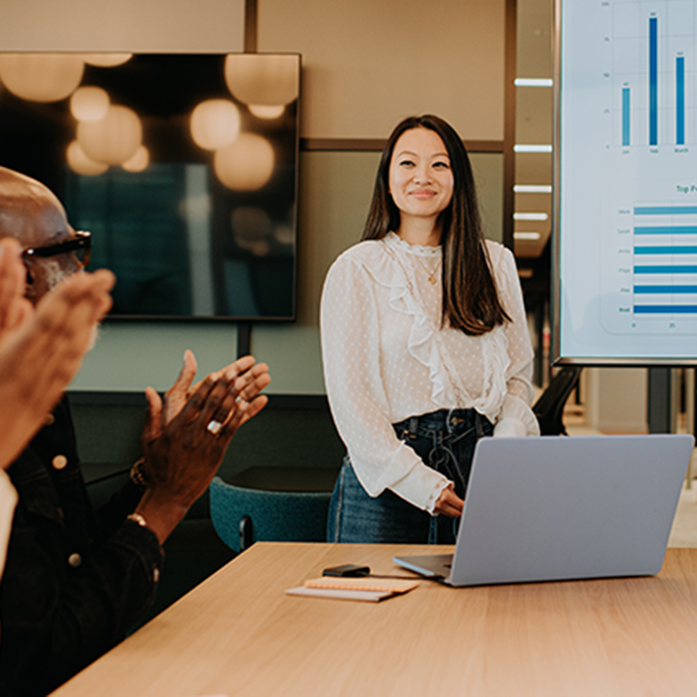 A businesswoman stands confidently in front of a large screen displaying charts and graphs. Colleagues applaud her presentation in a modern conference room.