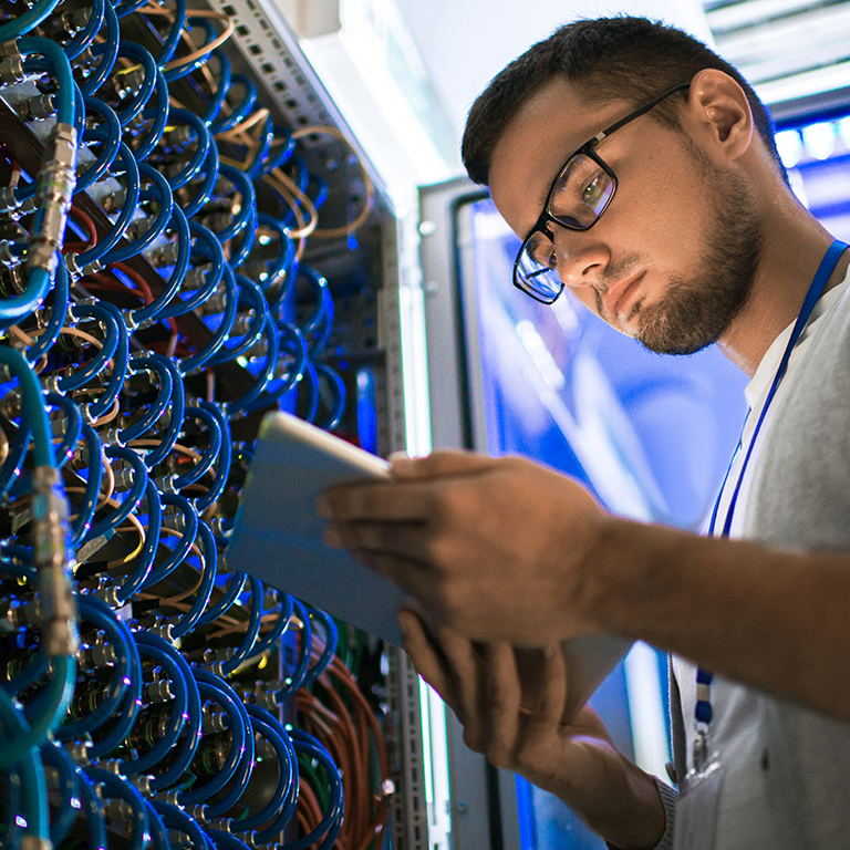 Low angle  portrait of young man using digital tablet standing by server cabinet while working with supercomputer in blue light