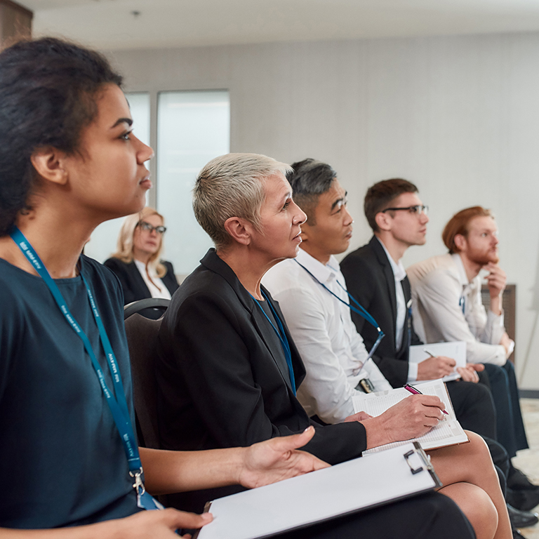 Multi-ethnic audience sitting in a row and making notes while listening to presentation at a business seminar. Selective focus. Horizontal shot. Side view