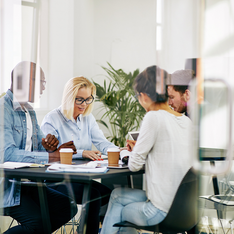 Diverse group of young businesspeople sitting around a table inside of a glass walled boardroom having a meeting together