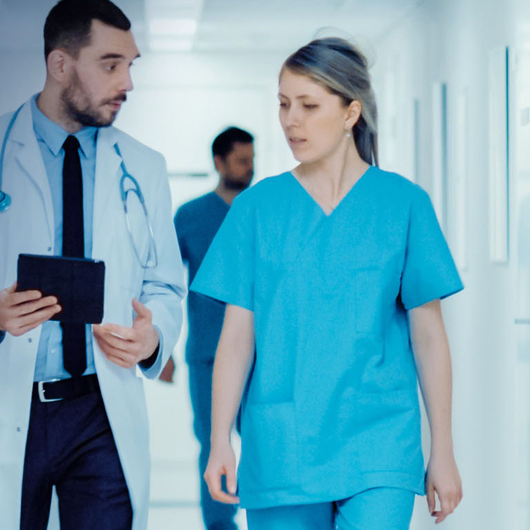 Surgeon and Female Doctor Walk Through Hospital Hallway, They Consult Digital Tablet Computer while Talking about Patient's Health. Modern Bright Hospital with Professional Staff. 