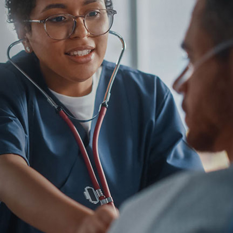 Hospital Ward: Friendly Black Head Nurse Uses Stethoscope to Listen to Heartbeat and Lungs of Recovering Male Patient Resting in Bed, Does Checkup. Man Getting well after Successful Surgery