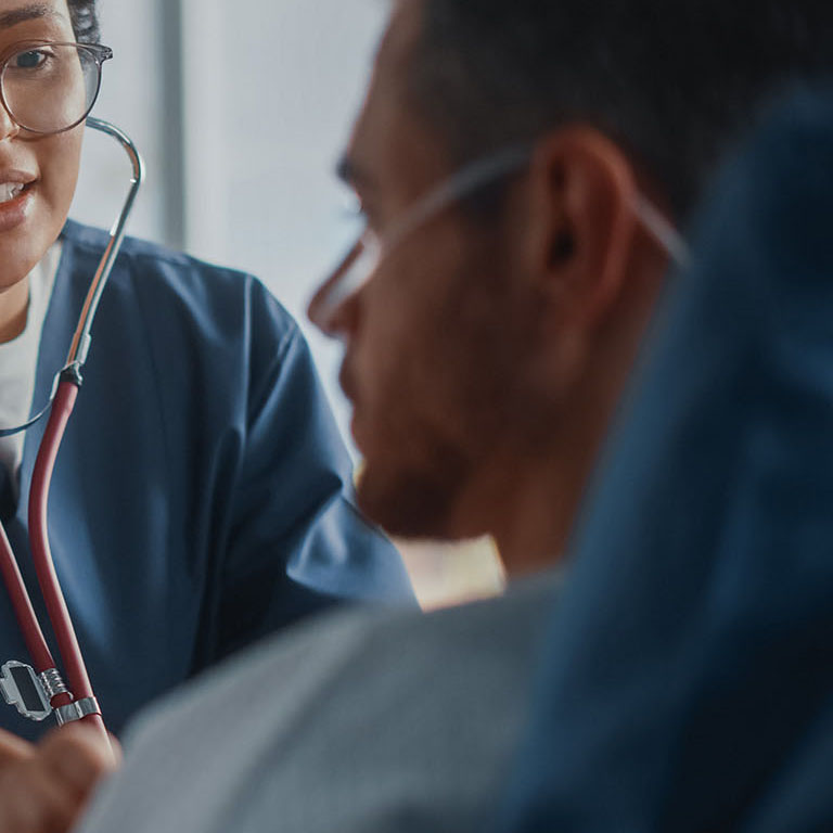 Hospital Ward: Friendly Black Head Nurse Uses Stethoscope to Listen to Heartbeat and Lungs of Recovering Male Patient Resting in Bed, Does Checkup. Man Getting well after Successful Surgery