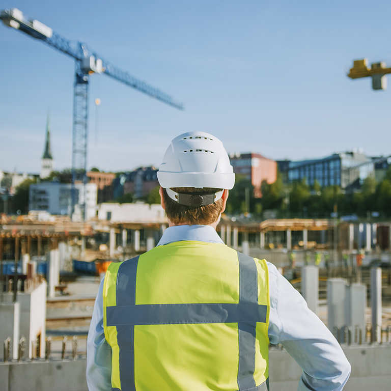 Confident bearded head Civil Engineer-Architect in sunglasses is Sstanding outside with his back to camera in a construction site on a bright day. Man is wearing a hard hat, shirt and a safety vest.