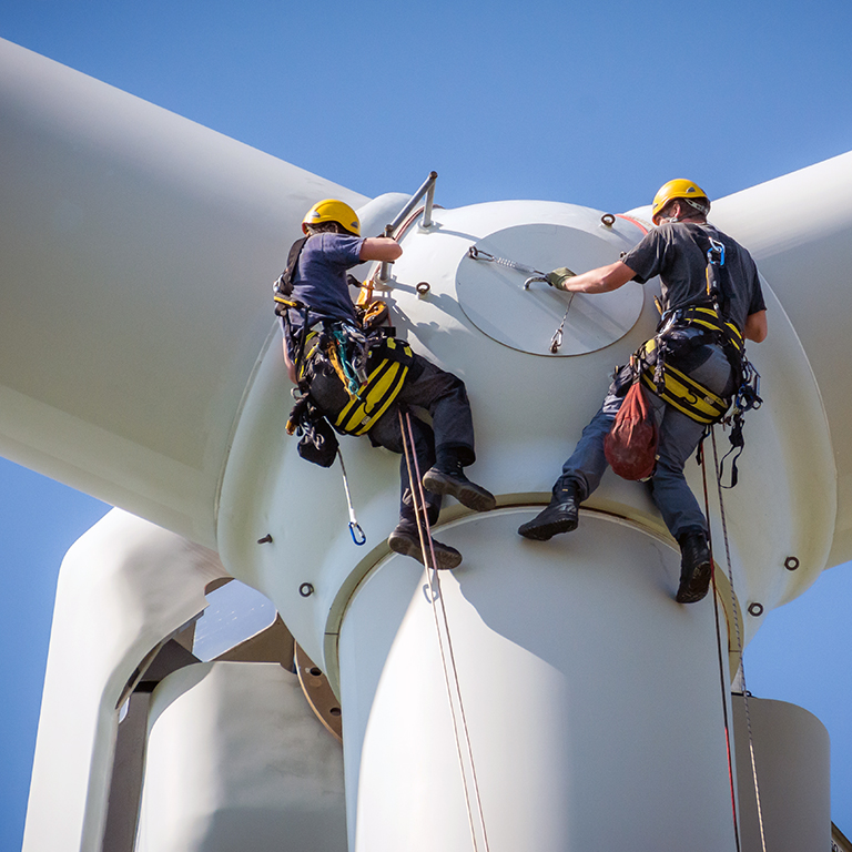 Inspection engineers preparing to rappel down a rotor blade of a wind turbine in a wind farm on a clear day with blue sky.