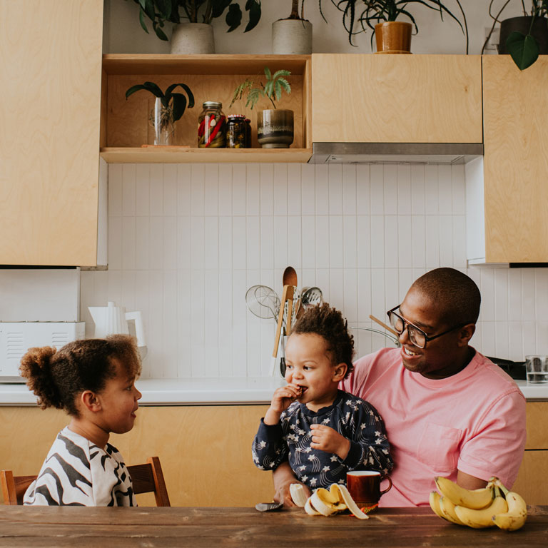 A father sits at a long dining table in a stylish domestic kitchen. One of his children sits on his lap, and the other sits on a chair. He talks to them as they enjoy a snack.