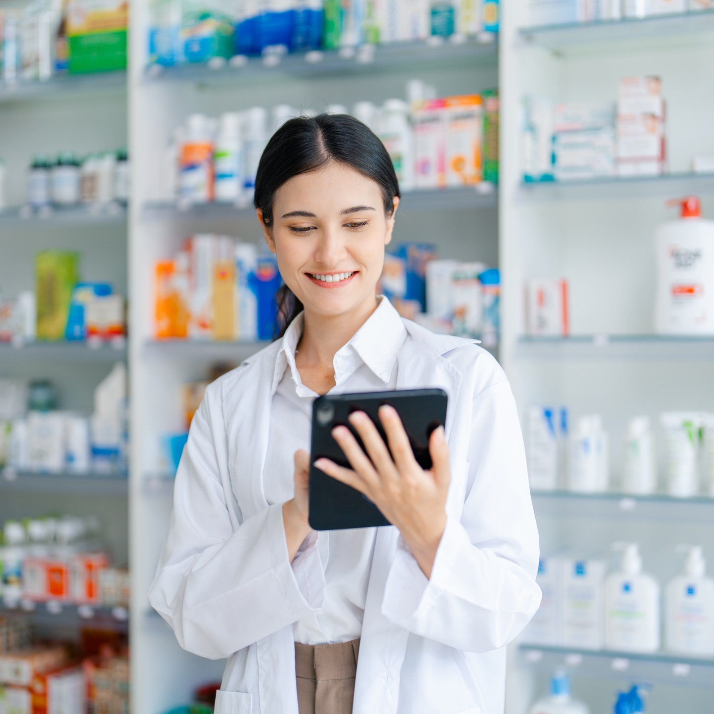 Female pharmacist in a white coat uses a digital tablet to assist with pharmacy services, showcasing the integration of modern technology in a well-stocked healthcare setting.