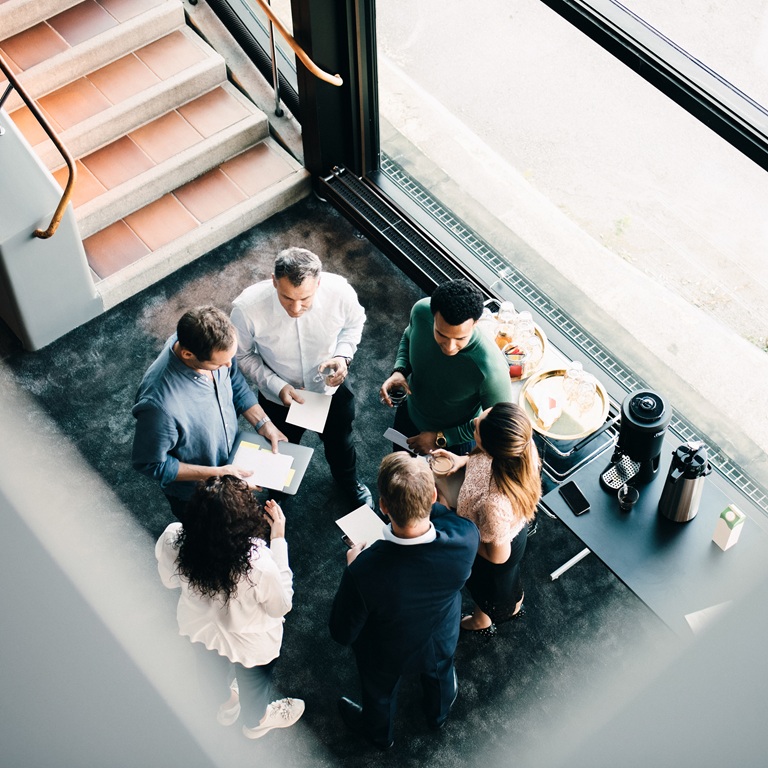 A group of people gathers in a modern building to discuss plans. They talk and share papers while standing near a coffee station by the windows.