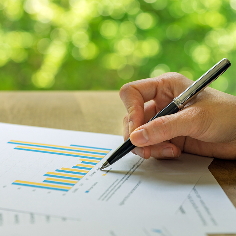 Business, investment or financial report review, hand holding pen reviewing bar graph information data print on paper with green bokeh in the background.