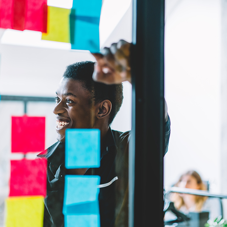 Cheerful African American men entrepreneur standing in front of wall with stickers and making notes of ideas for startup management project, happy smiling male employee working in modern office