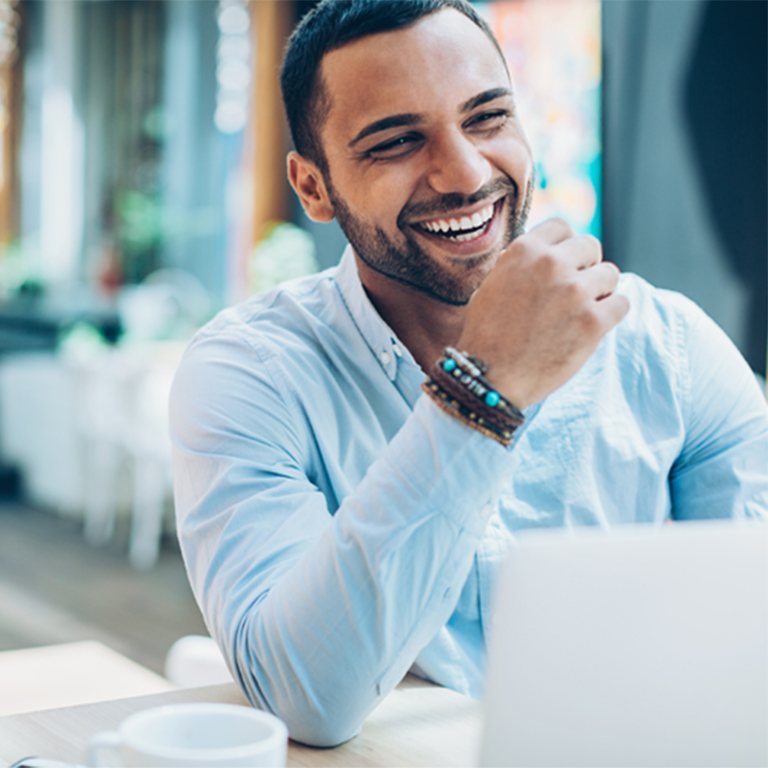 A man is sitting at a table in a bright café, smiling and interacting with his laptop. He enjoys a drink, showcasing a relaxed and productive atmosphere during the day.
