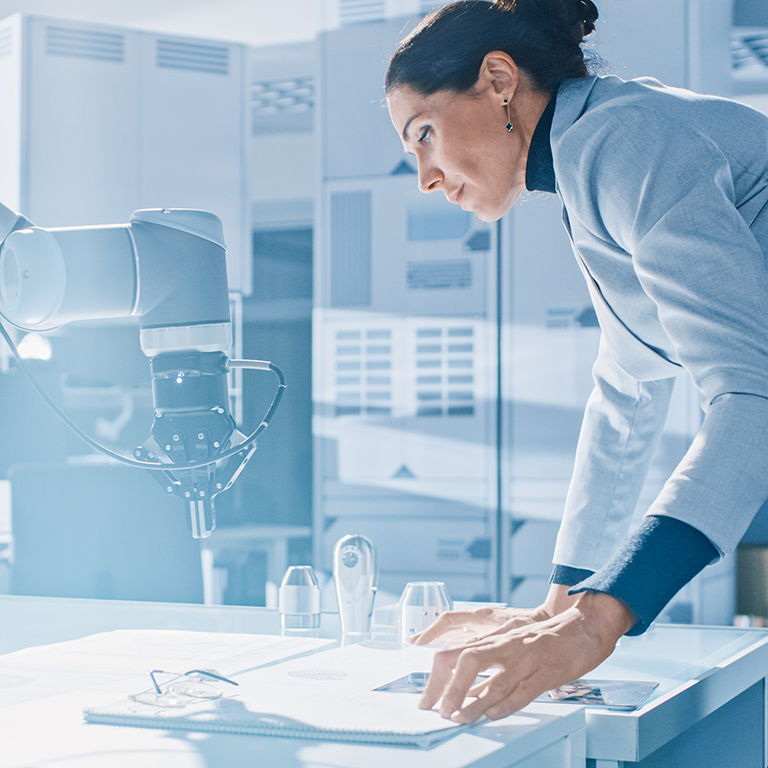 Stylish Female Robotics Engineer Leans on the Table Works with Blueprints, Documents and Tablet Computer, She's Programming Robot Arm Movements