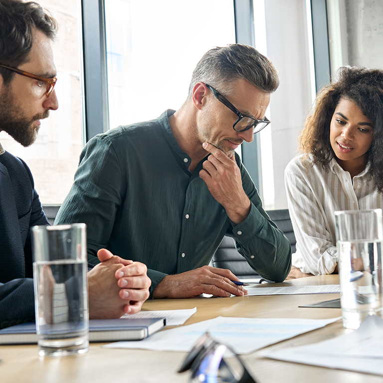 Focused doubtful mature businessman reading contract document thinking considering risks with professional experts executive team analyzing financial report sitting at office table.