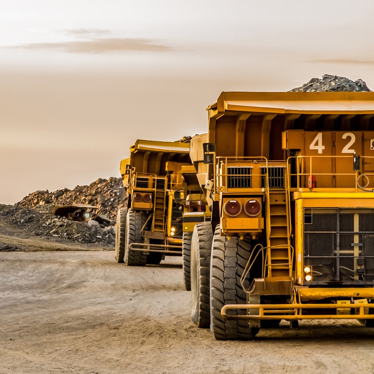 Two massive mining trucks drive along a dirt road amid a rocky landscape as the sun sets, casting a warm glow over the scene. Dust rises from their tires, highlighting their large size.