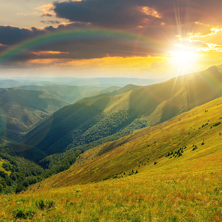 mountain landscape in summer at sunset. grassy meadows on the hills rolling in to the distant peak beneath a rainbow in evening light