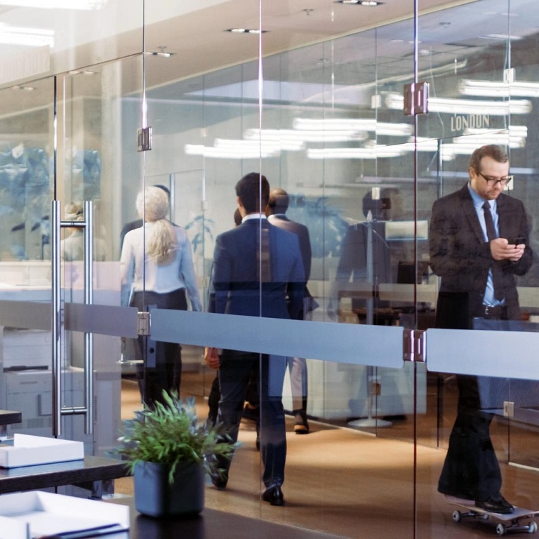 Suited Asian Businessman Rides Skateboard Through the Corporate Building Hallway. Stylish Glass and Concrete Building with Multicultural Crowd of Business People.