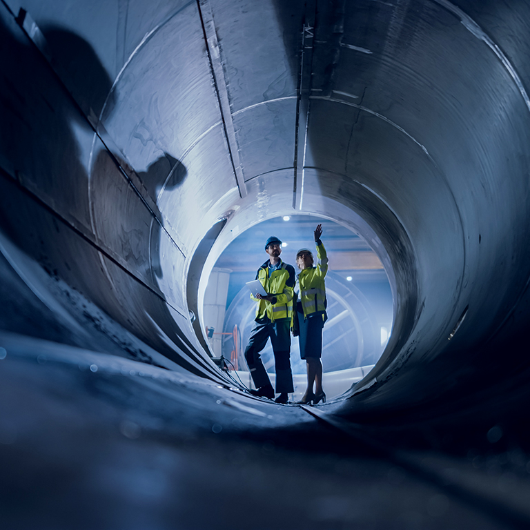 Two Heavy Industry Engineers Walking Inside Pipe, Use Laptop, Have Discussion, Checking Design. Construction of the Oil, Natural Gas and Biofuels Transport Pipeline. Industrial Manufacturing Factory