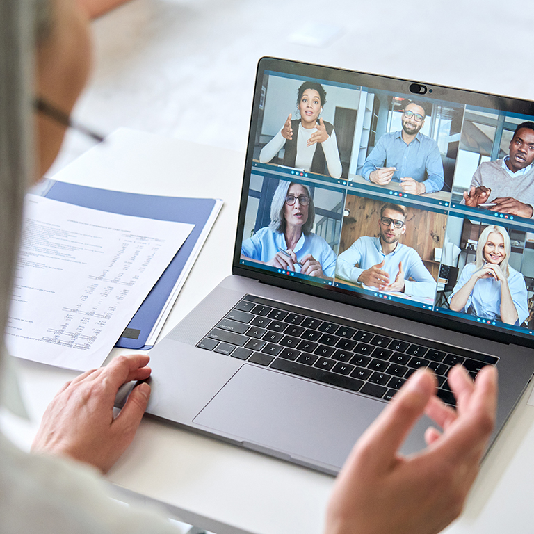 Asian mid age businesswoman ceo executive manager in headset having working videocall on financial report with multiracial colleagues using laptop in modern corporation office. Over shoulder view.