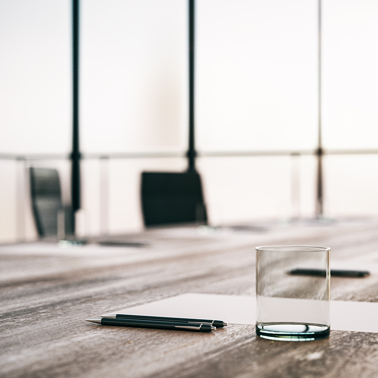 Close up of wooden boardroom table with glass, pens and blank sheet at blurry window background in the morning. 