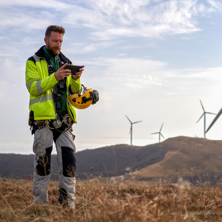 Renewables worker in hi vis checking a tablet in a field with wind turbines in the background