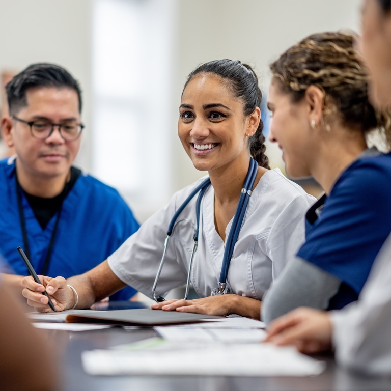 A small medical staff team sit around a boardroom table as they meet to discuss and collaborate on patient cases.  They are each dressed professionally and are focused on the meeting.