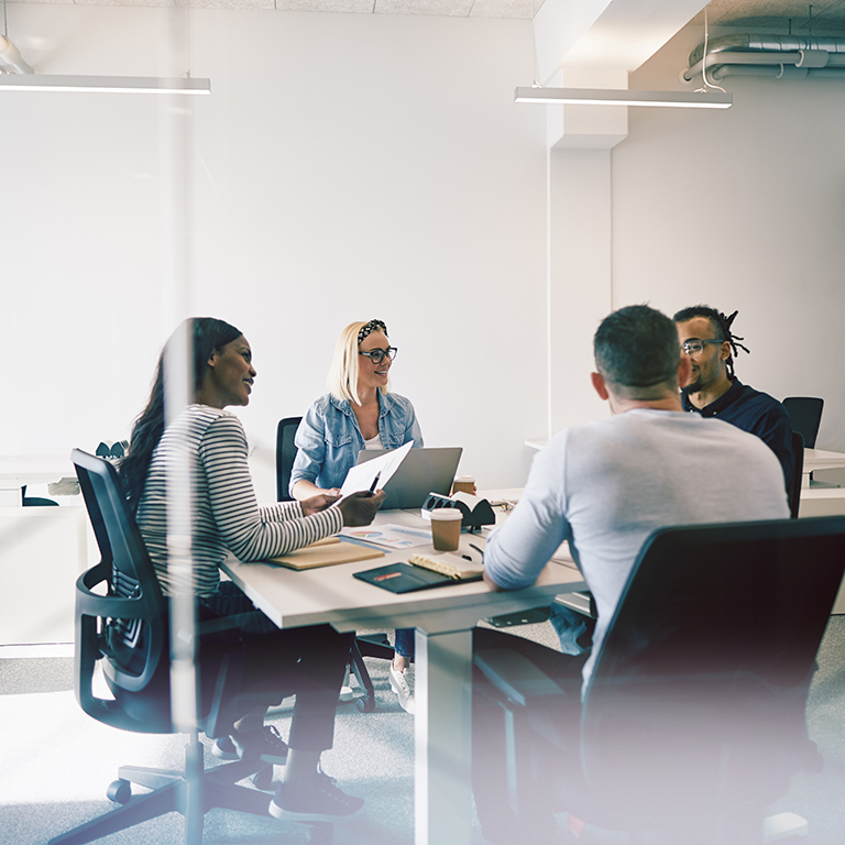 Smiling group of diverse coworkers talking together during a meeting around a table inside of a glass walled office