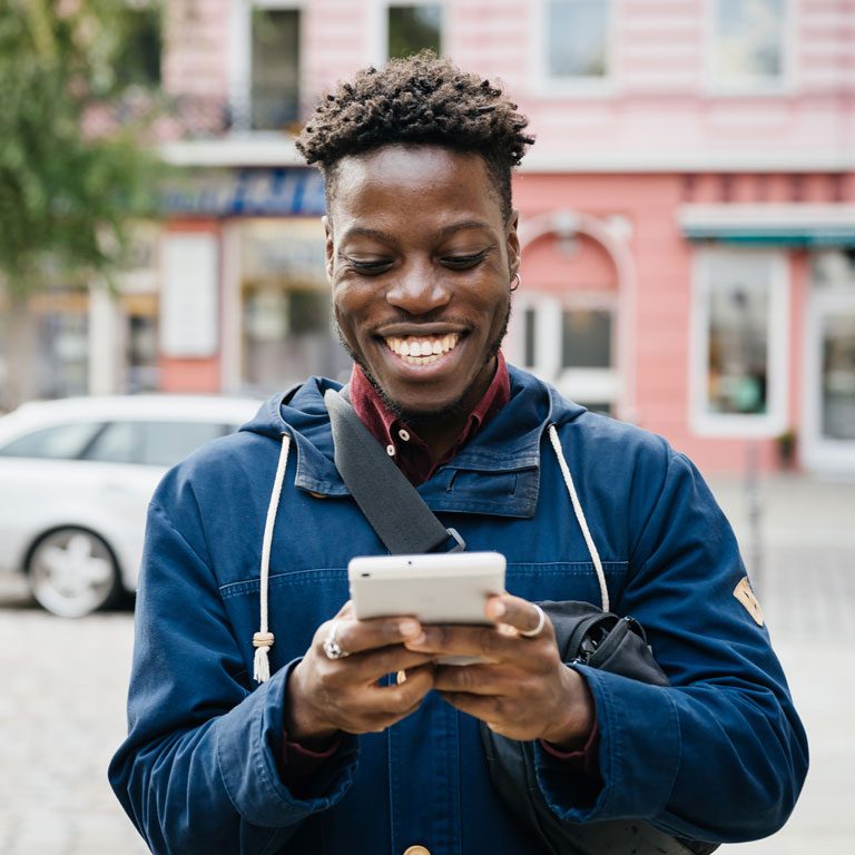 A man smiling while using his smartphone while out in the city.