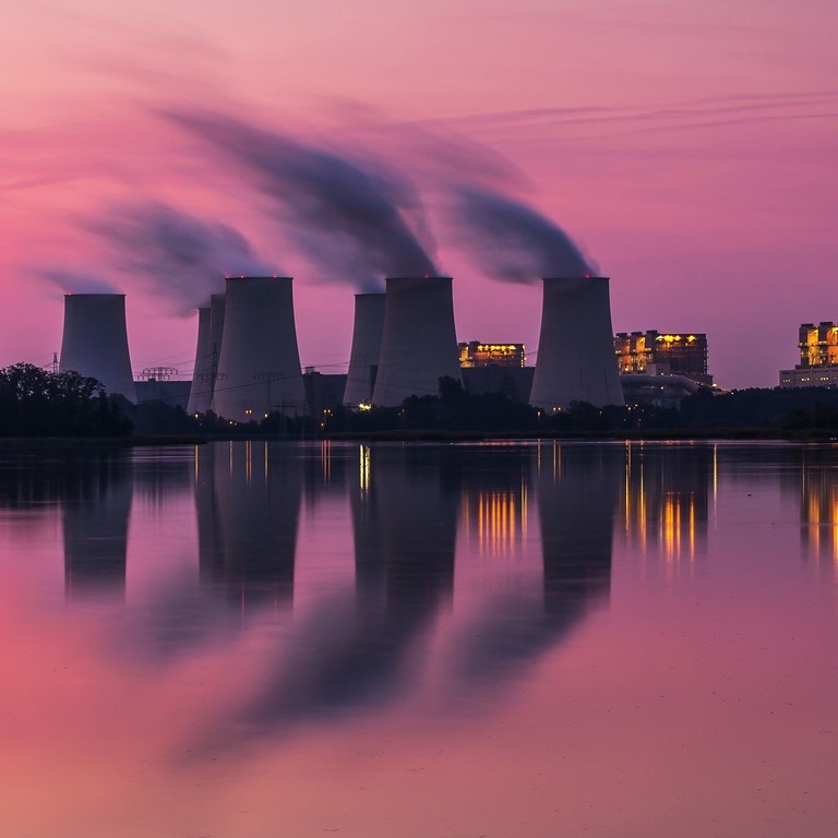 Smoke billows from cooling towers at a power plant during sunset. The purple sky reflects on the calm water below. Lights from the plant shine in the dusk.