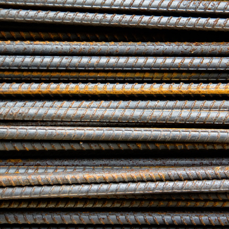 Steel bars are stacked closely together at a construction site. They show signs of wear and rust. The setting is likely in an urban area during daylight hours.
