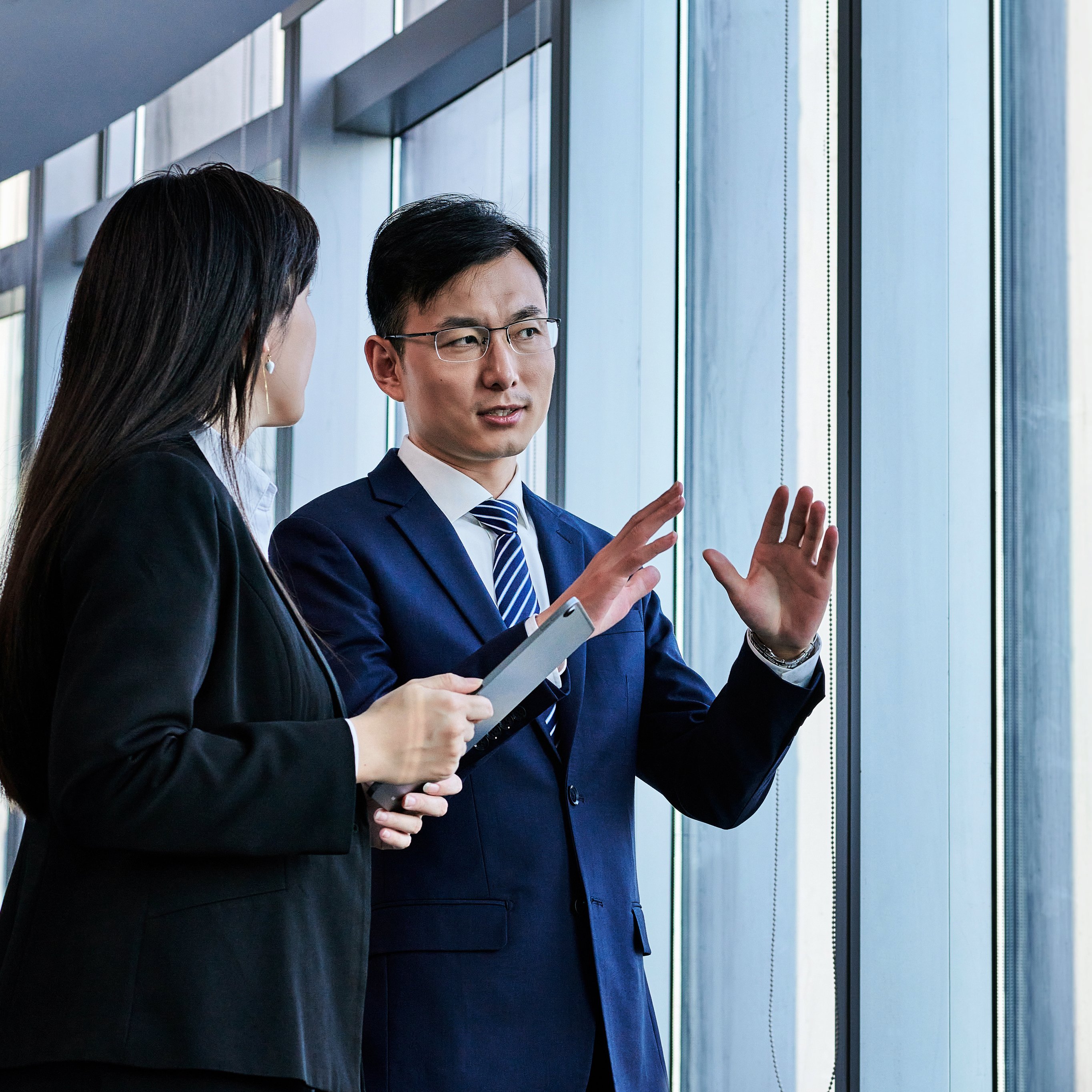 Two professionals stand by a large window in an office while discussing ideas and plans for work. The atmosphere shows focus and engagement.