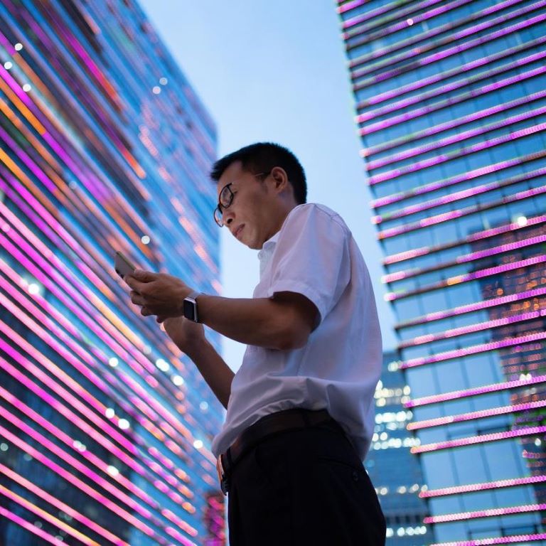 A man in a white shirt stands focused on his phone, surrounded by vibrant, illuminated structures as twilight descends, highlighting the modern urban landscape.